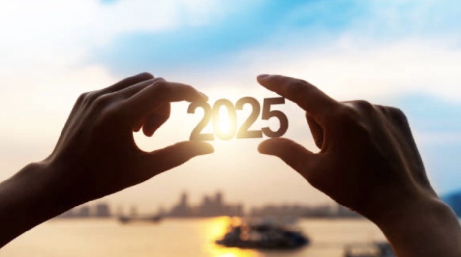 A pair of hands are holding up the number 2025 with the sky in the background. The sun shines through the numbers, highlighting the year. Photo courtesy of Flickr.